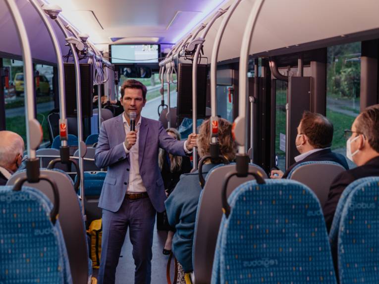 Ein Herr steht in einem Bus und spricht zu den anderen Mitfahrenden in ein Mikrofon